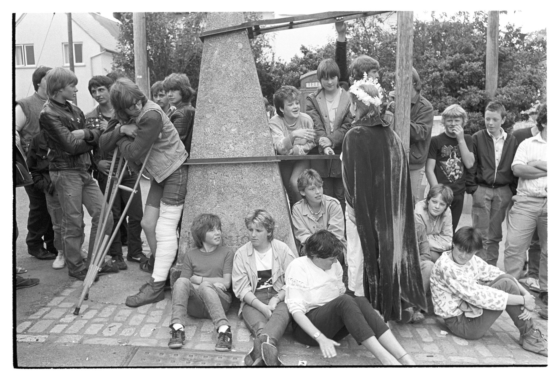 Crowd of young people around village pump at start of Fair. 
[A group of young men and women stood and sat around the village pump at the start of Winkleigh Fair. The Fair Queen is talking to some of the people and one young man has a pair of crutches.]