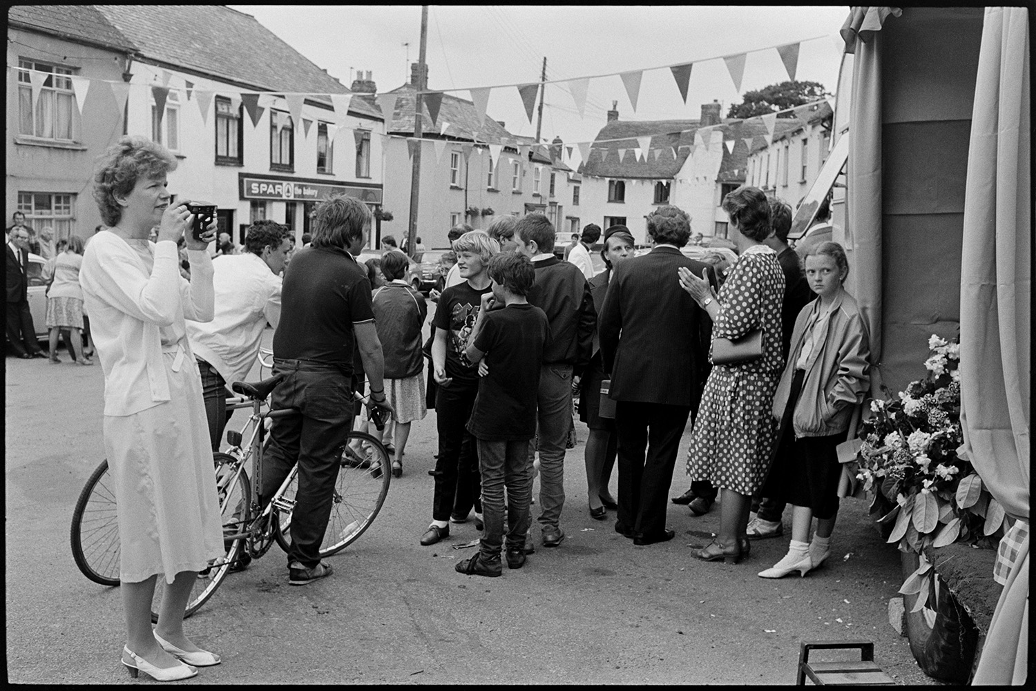 Fair Queen and attendants in village square, spectators waiting around. Photographer. 
[A woman taking a photograph of a caravan which has been converted into a stage at Winkleigh Fair. Other people are gathered around, including children and two teenagers with bicycles. The spar shop can be seen in the background and the street is decorated with bunting.]