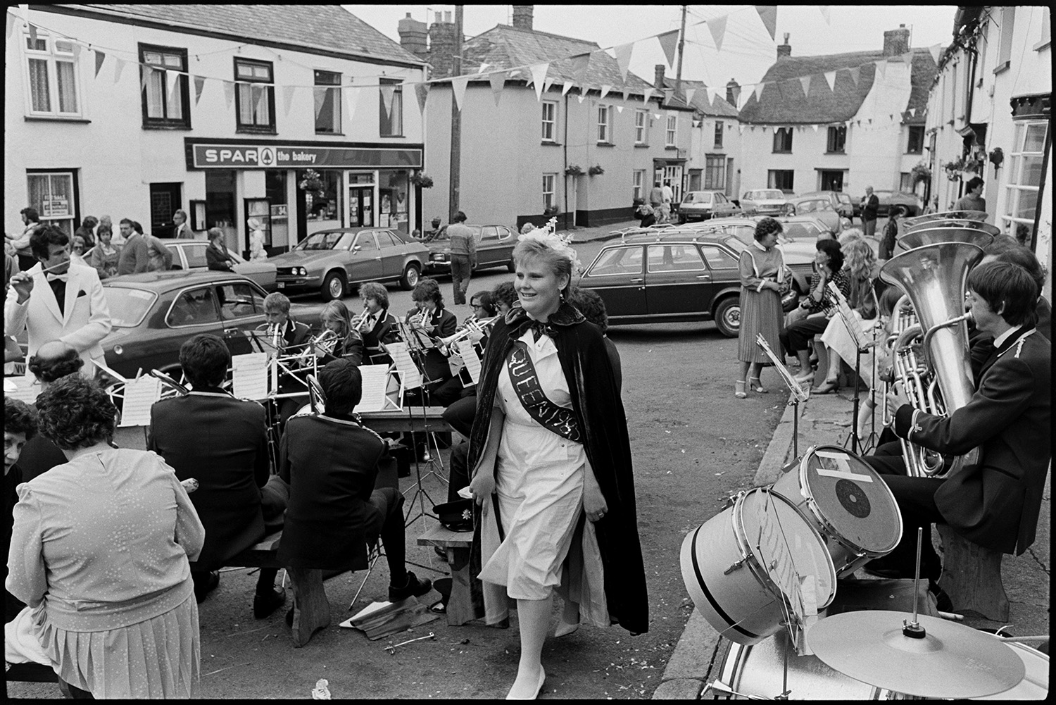 Fair Queen and attendants in village square, spectators waiting around. Photographer. 
[The Winkleigh Fair Queen walking through Winkleigh village square past a bras band. The conductor of the band is visible in the background. Thatched cottages, parked cars and the Spar shop are visible in the street which is decorated with bunting.]