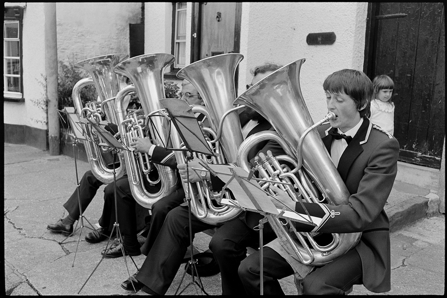 Tuba players of brass band playing at village fair. 
[Four tuba players performing with a brass band at Winkleigh Fair. They are sat outside a cottage in Winkleigh square.]