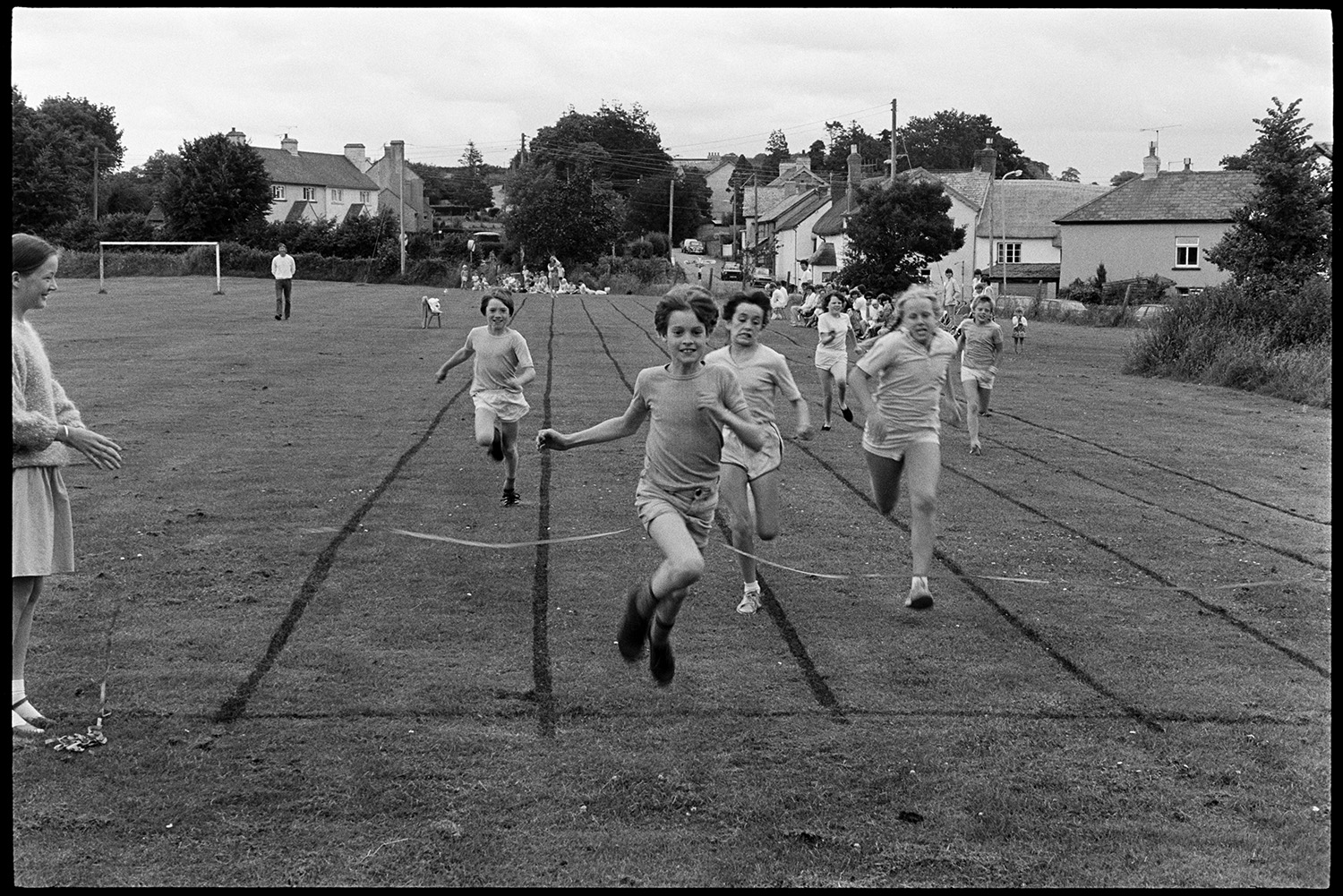 School Sports day, races, sack race, egg and spoon. 
[Children competing in a running race at Dolton School sports day. A boy is crossing the finishing line and winning the race. Spectators are watching in the background.]