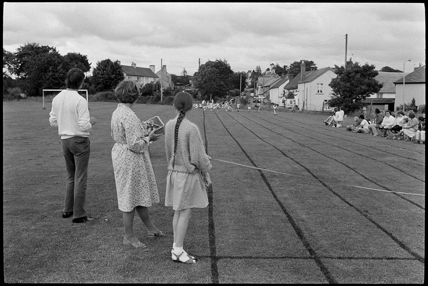 School Sports day, races, sack race, egg and spoon. 
[A man, woman and girl stood at the finish line at of the track at Dolton School sports day, judging the races. The girl is holding the rope across the finish line. In the background children are lining up for a race and spectators are watching from the side of the track.]
