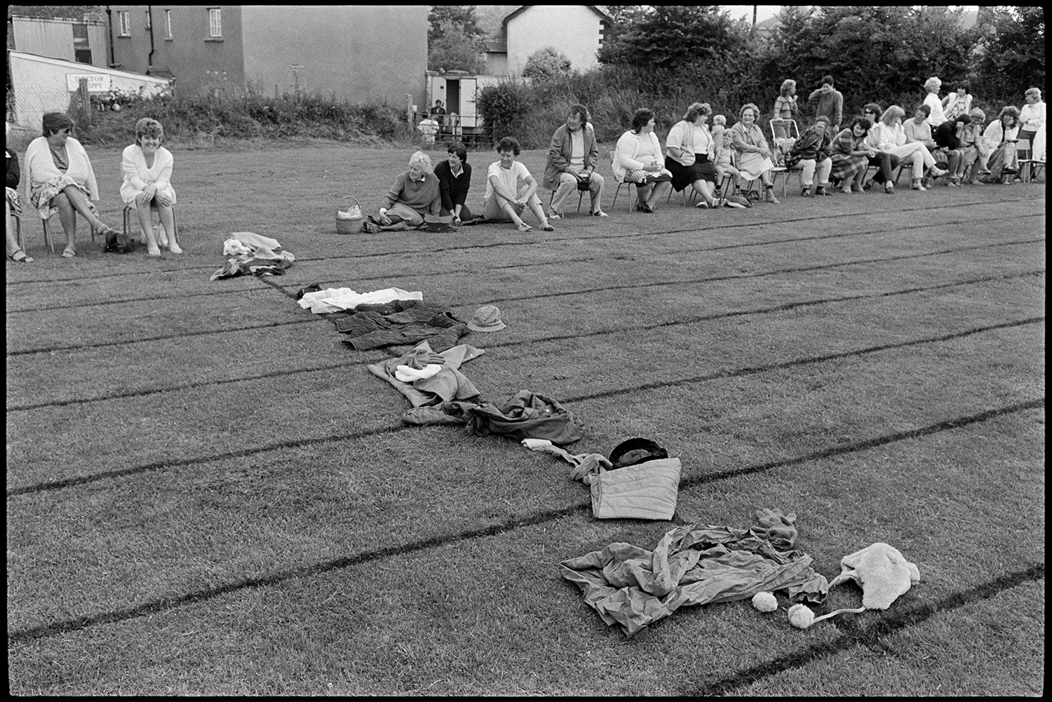 School Sports day, races, sack race, egg and spoon. 
[Clothes lines up on the race track at Dolton School sports day for a race. Spectators, including parents, are sat along the side of the track to watch the races.]