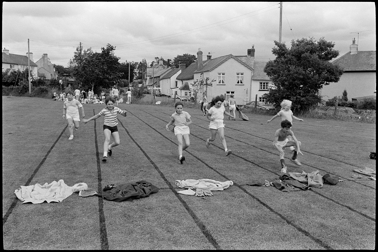 School Sports day, races, sack race, egg and spoon. 
[Children competing in a clothes race at Dolton School sports day. Other children are watching from the other end of the track and houses can be seen in the background.]