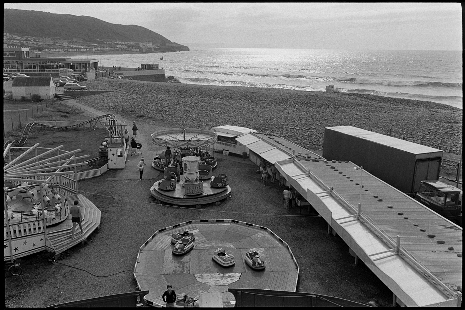 Seaside amusements, children. 
[An amusement park on Westward Ho! beach with rides. A small rollercoster, merry-go-round and dodgems are visible on the seafront.]