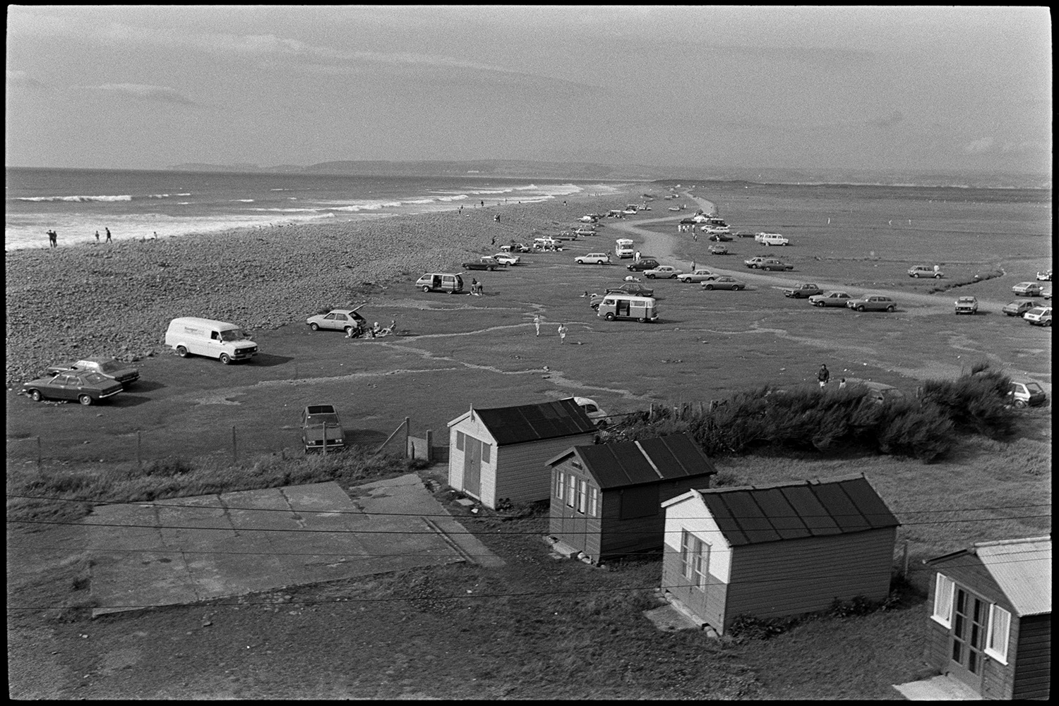 Seaside amusements, children. 
[Cars parked behind the pebble ridge at Westward Ho! beach. People are walking along the beach and beach huts can be seen in the foreground.]