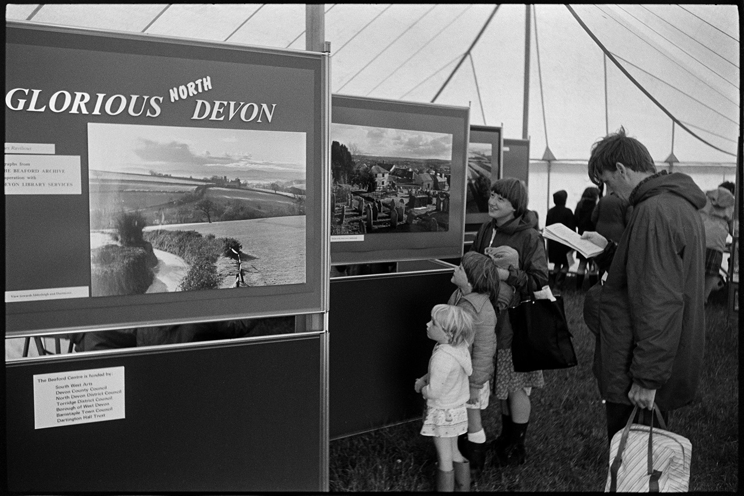 North Devon Show, prize bulls and cattle. 
[A man, woman and children looking at a display of images from the Beaford Archive, taken by James Ravilious in a tent at the North Devon Show at Alverdiscott.]