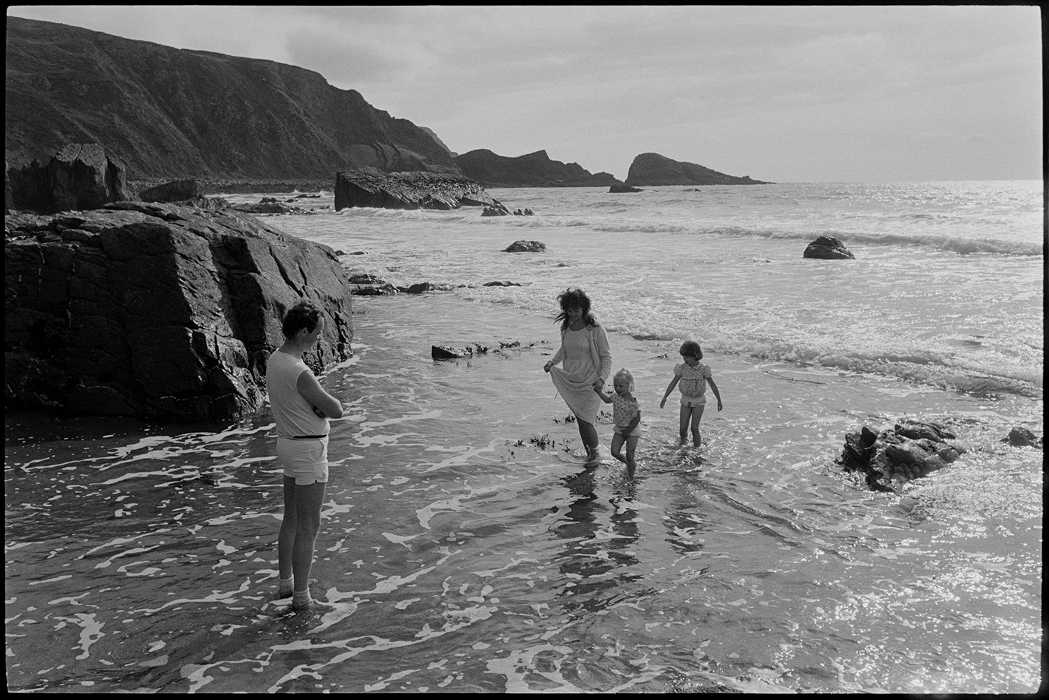 Family on seashore. 
[Carol Duncan and Peter Duncan paddling in the sea on the beach at Welcombe, with their children. The seashore and cliffs can be seen in the background.]