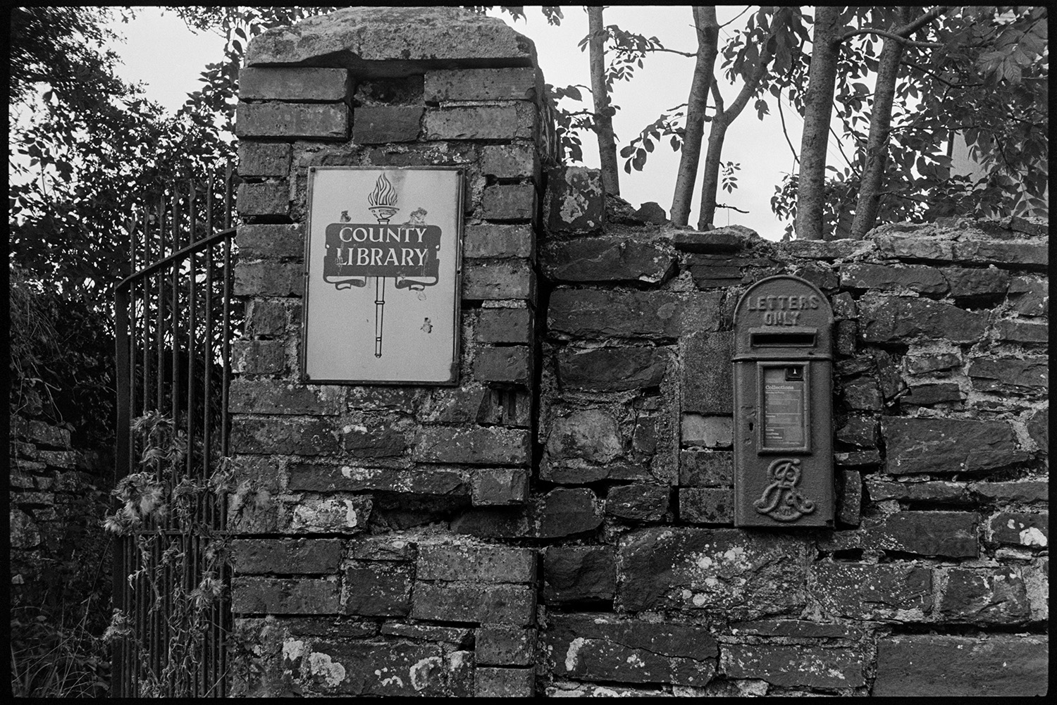 Letter box in wall, old. 
[A post box set into a stone and brick wall by a gateway with a sign for a Country Library.]