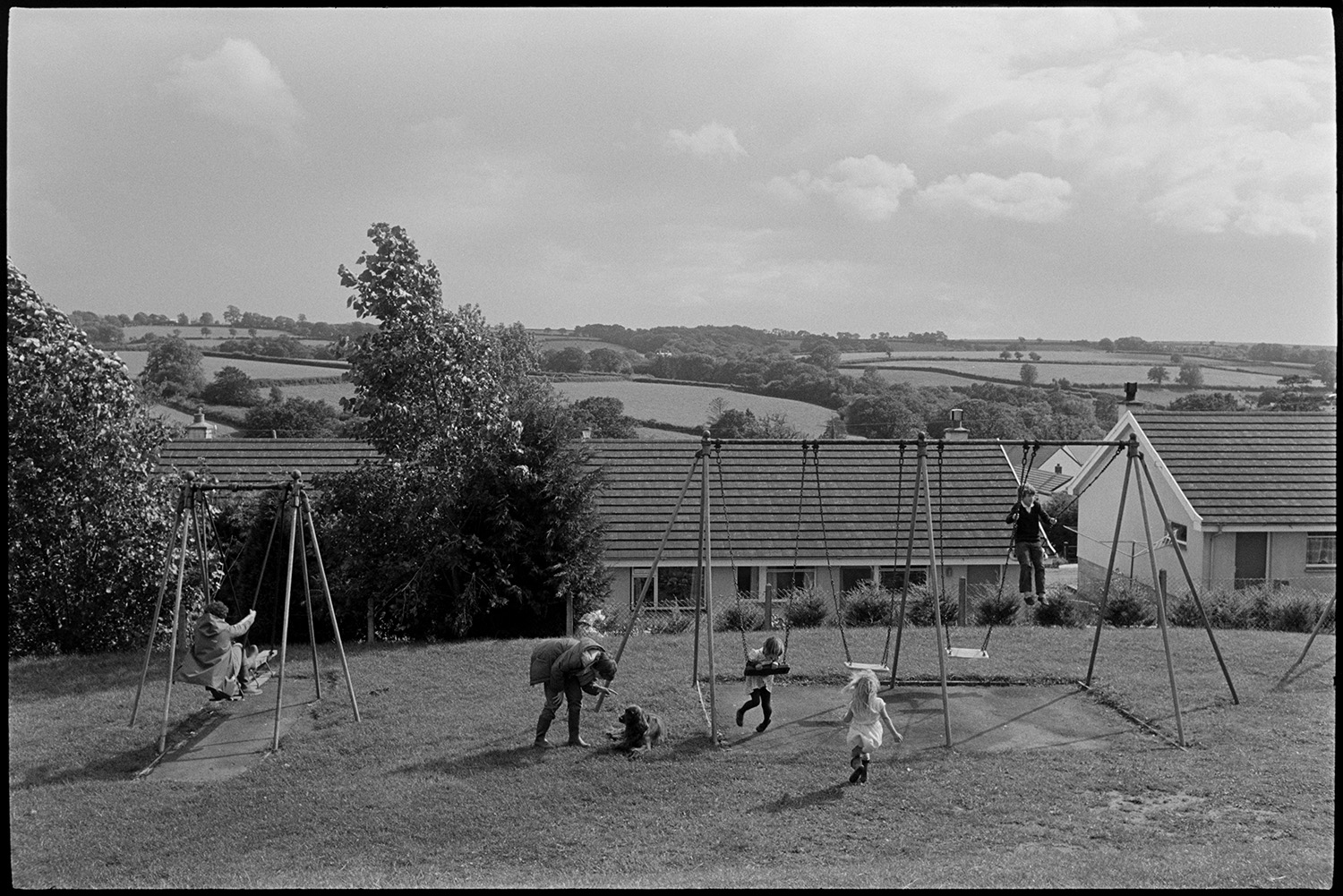 Children playing on swings in public playground. 
[Children playing on swings in a playground at Stafford Way, Dolton. A person is telling off a dog by the swings. Houses, fields and trees can be seen in the background.]