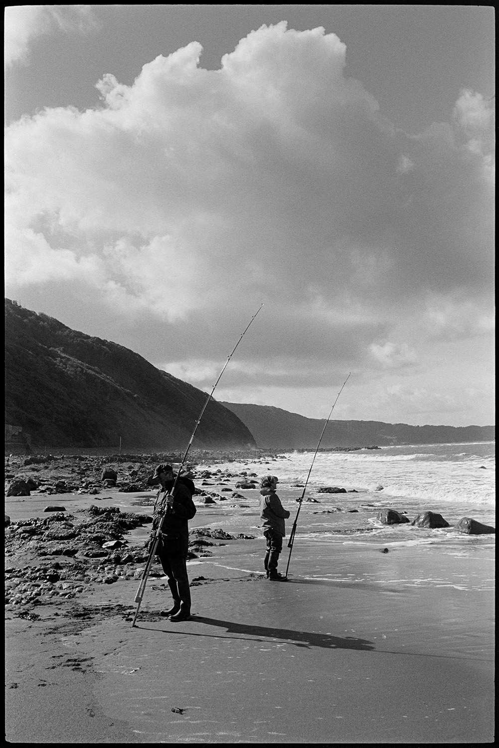 Seaside, beach with shells on rocks, limpets. 
[Two people fishing with fishing rods on the shoreline at Bucks Mills. Rocks can be seen on the beach and cliffs are visible in the background.]