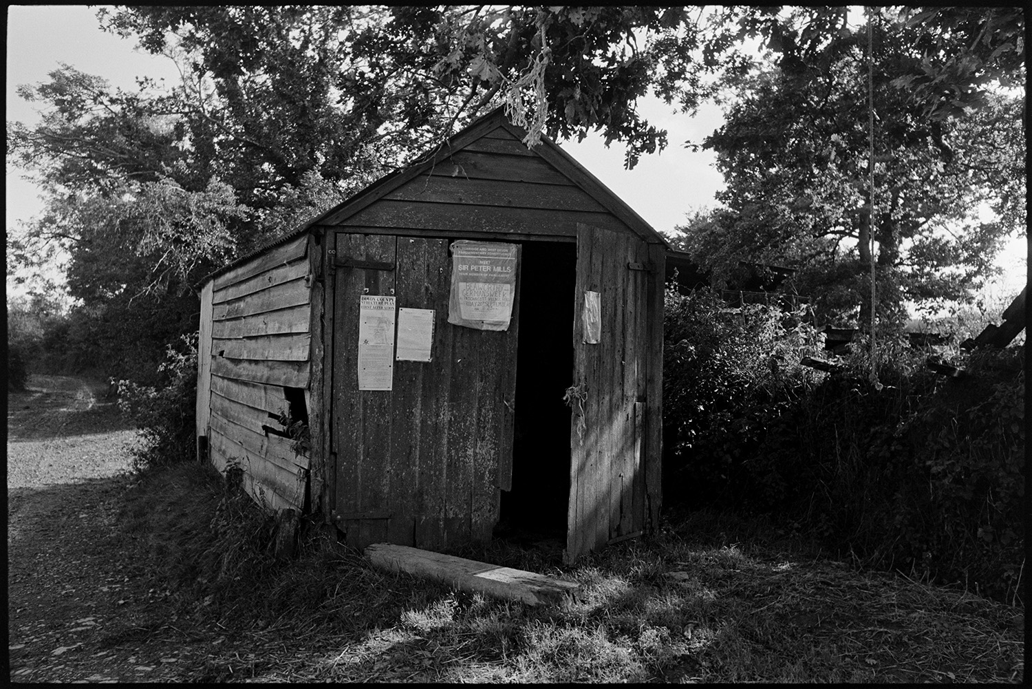 Small wooden shed, garage and house covered in posters. 
[A wooden shed or garage covered with posters, including one advertising a meeting with Sir Peter Mills MP, possibly near Northlew, Beaworthy.]