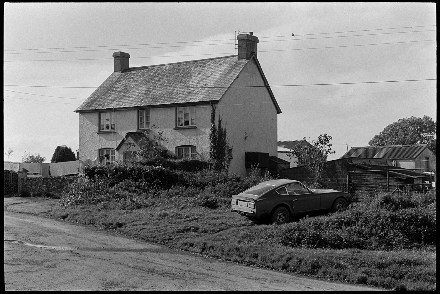 Small wooden shed, garage and house covered in posters. 
[A car parked outside a farmhouse possibly near Northlew, Beaworthy. Washing is hung out to dry on a washing line in front of the house and various sheds or barns can be seen in the background.]