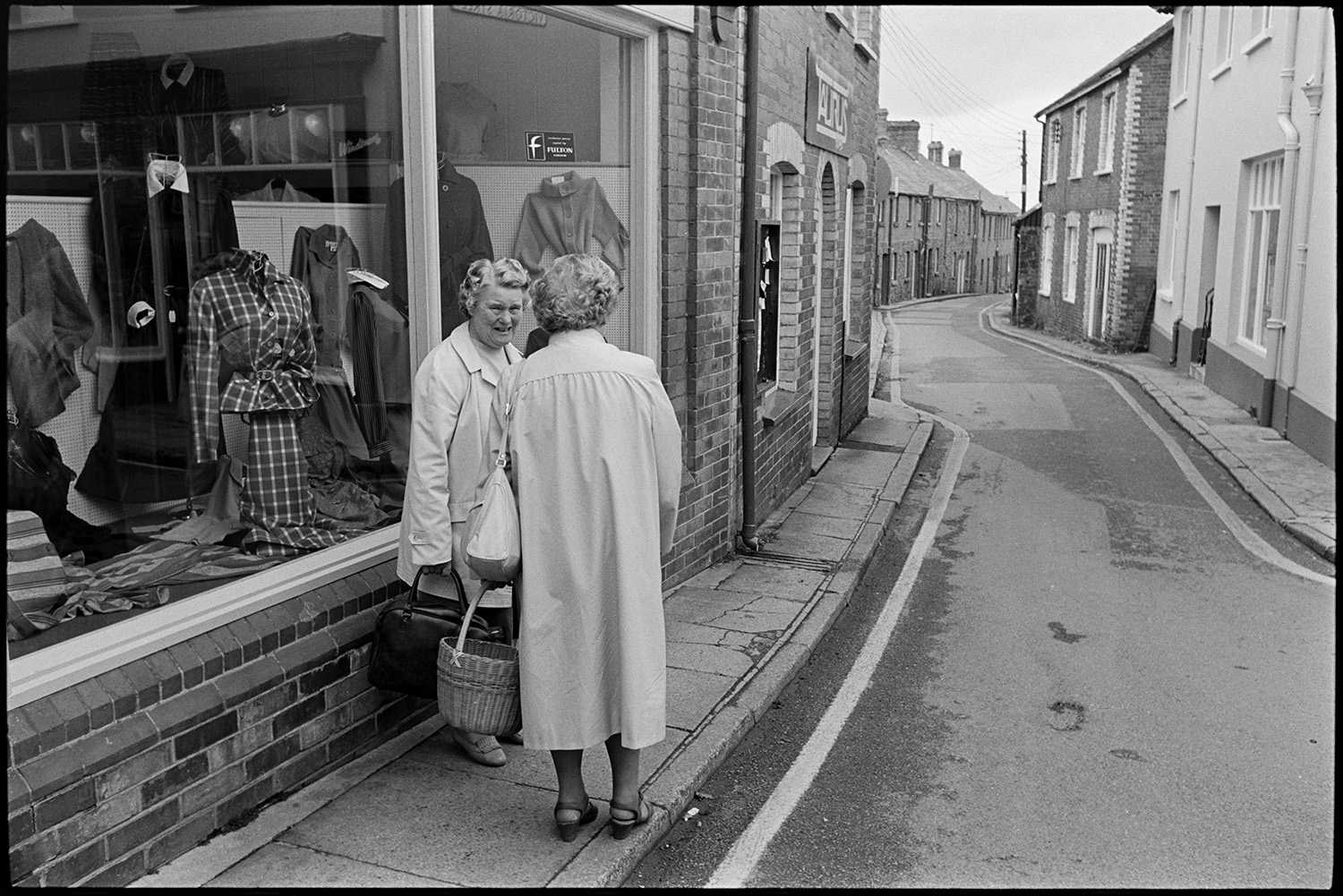 Town streets on market day clothes shop, people chatting. 
[Two women talking outside a clothes shop front window in Holsworthy. Terraced houses can be seen further along the street.]