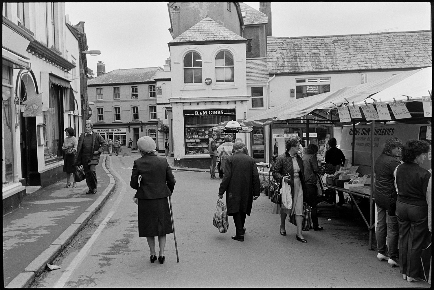 Town streets on market day clothes shop, people chatting. 
[Men and women shopping at market stalls in Holsworthy. Shop fronts are visible in the background including Headons Bakery and R&M Gibbs.]