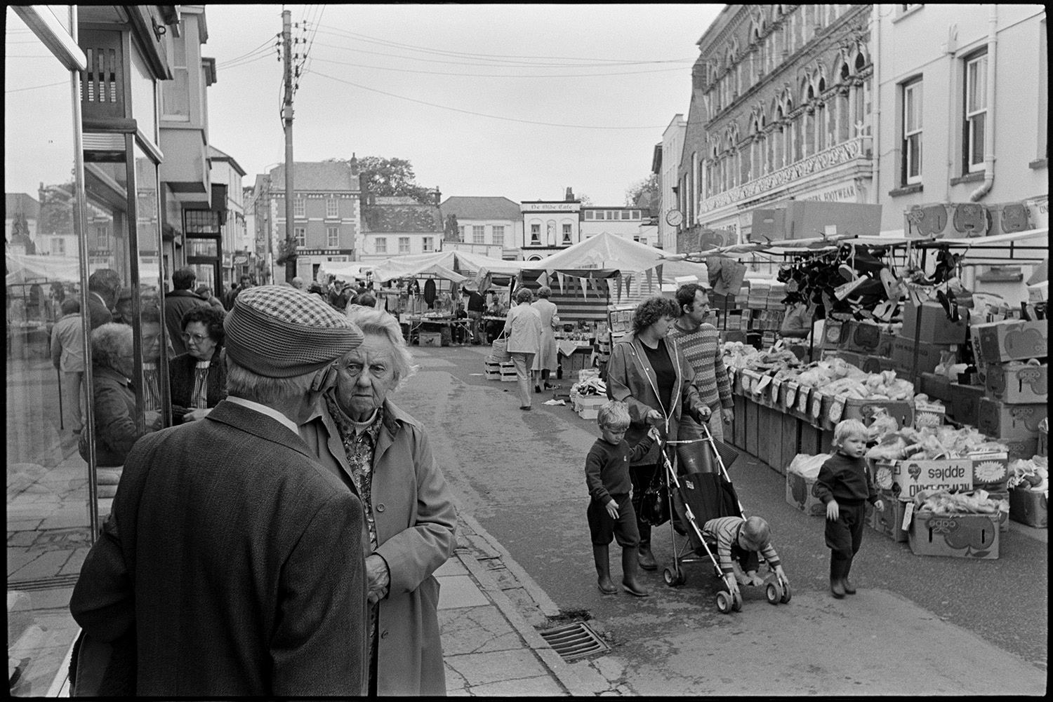 Town streets on market day clothes shop, people chatting. 
[People shopping in Holsworthy market. A family are walking past a stall selling shoes and a man and woman are talking in the foreground. Other market stalls can be seen in the background.]