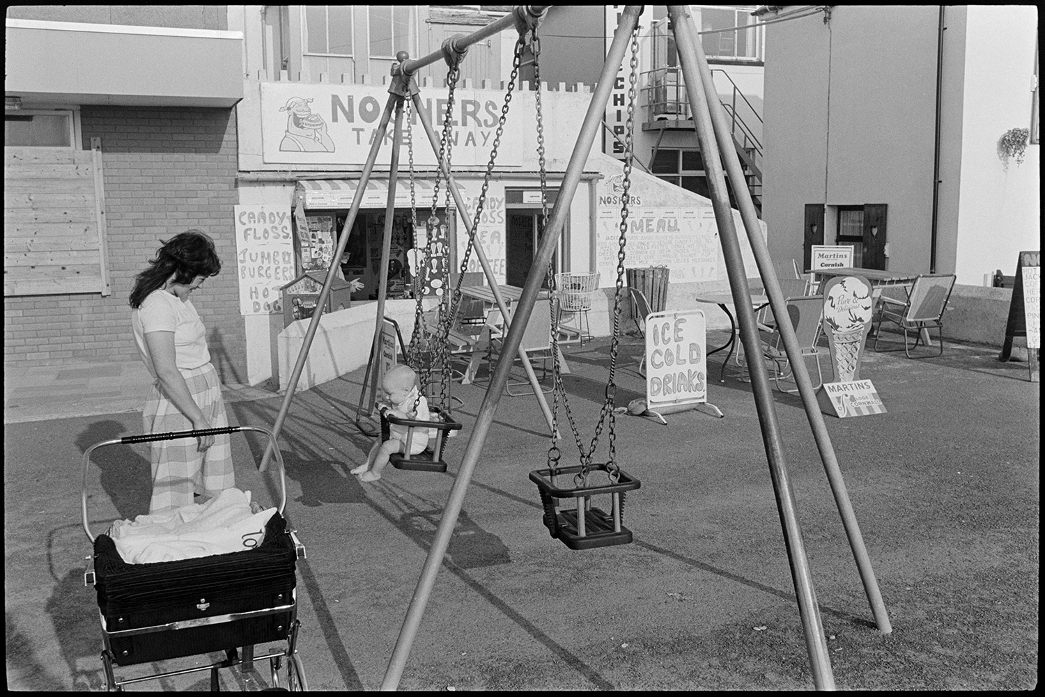 Mother and child playing on swing at seaside playground. 
[A mother pushing her baby on a swing in a playground outside a café at Westward Ho!. A pram is parked next to the swings.]