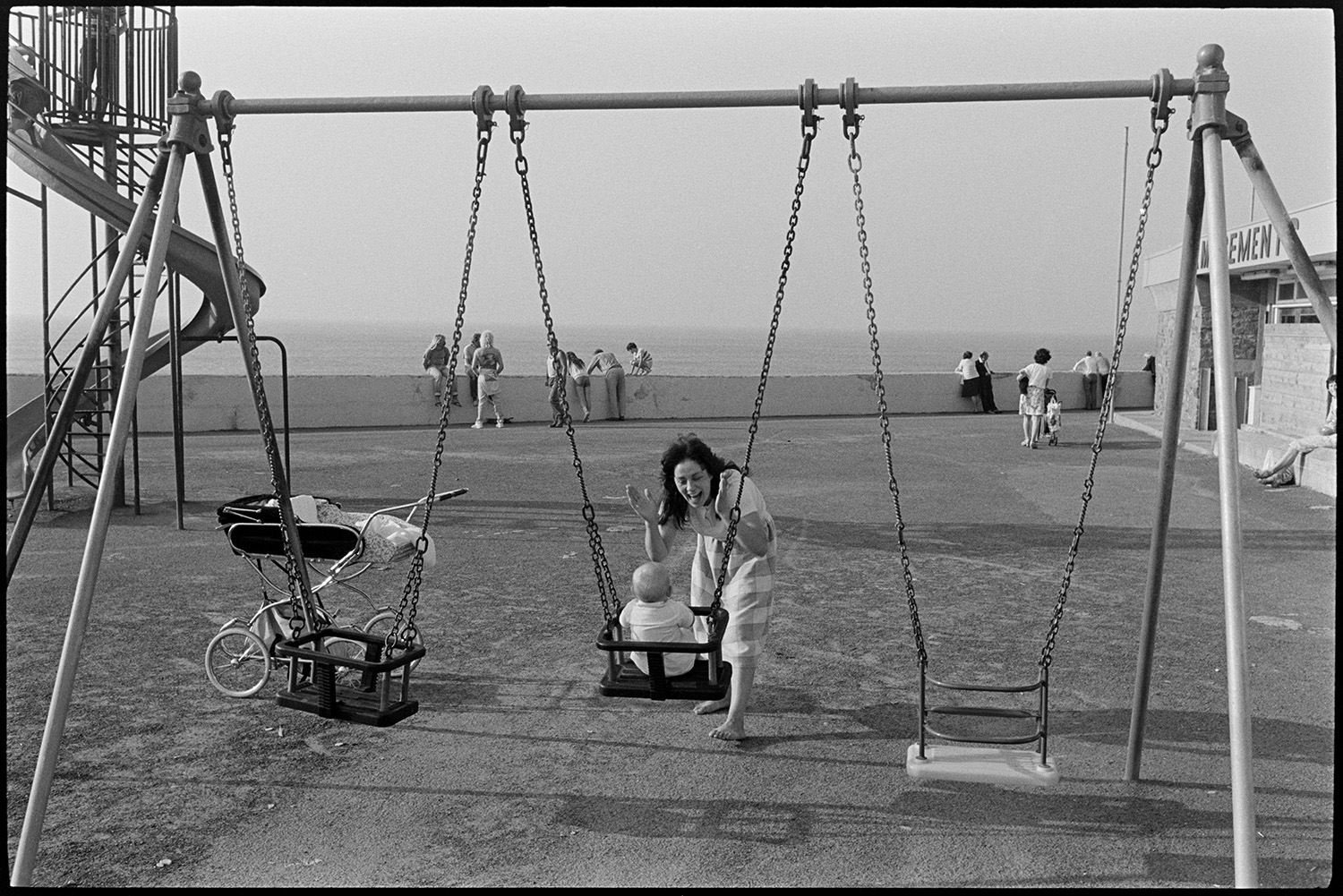 Mother and child playing on swing at seaside playground. 
[A mother pushing her baby on a swing in a playground outside a café at Westward Ho! seafront. A pram is parked next to the swings and people are sat on the sea wall in the background.]