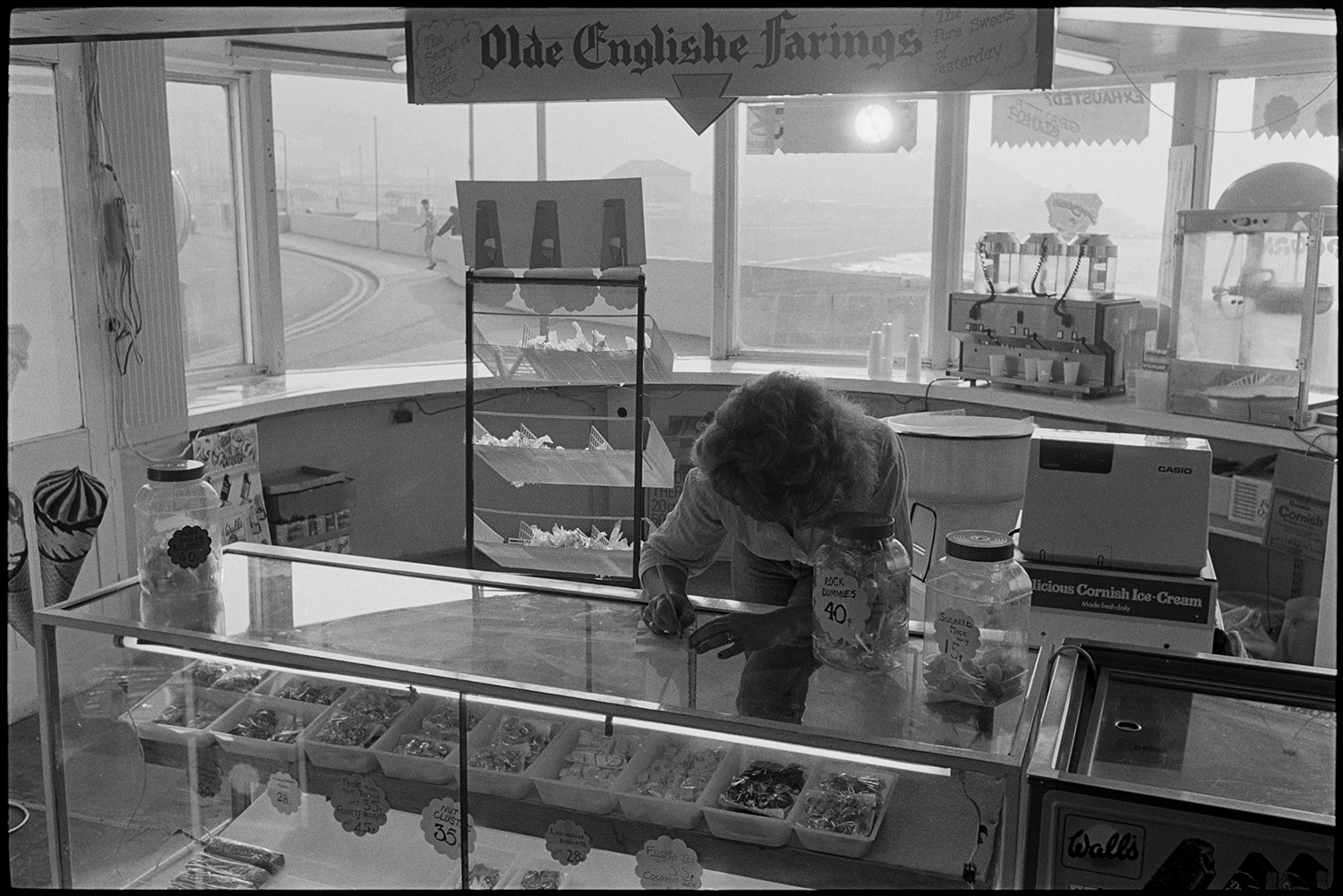 Counter of seaside sweetshop. 
[A woman writing a note on the counter of a sweet shop at Westward Ho!. Sweets are displayed in the glass counter and a fridge with ice cream is next to the counter. A till and two jars of sweets are on top of the shop counter.]