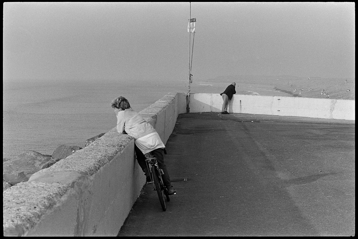 Boys leaning on sea wall looking at sea. 
[A boy on a bicycle leaning on the sea wall at Westward Ho! and looking out to sea. A man is also leaning on the sea wall in the background.]