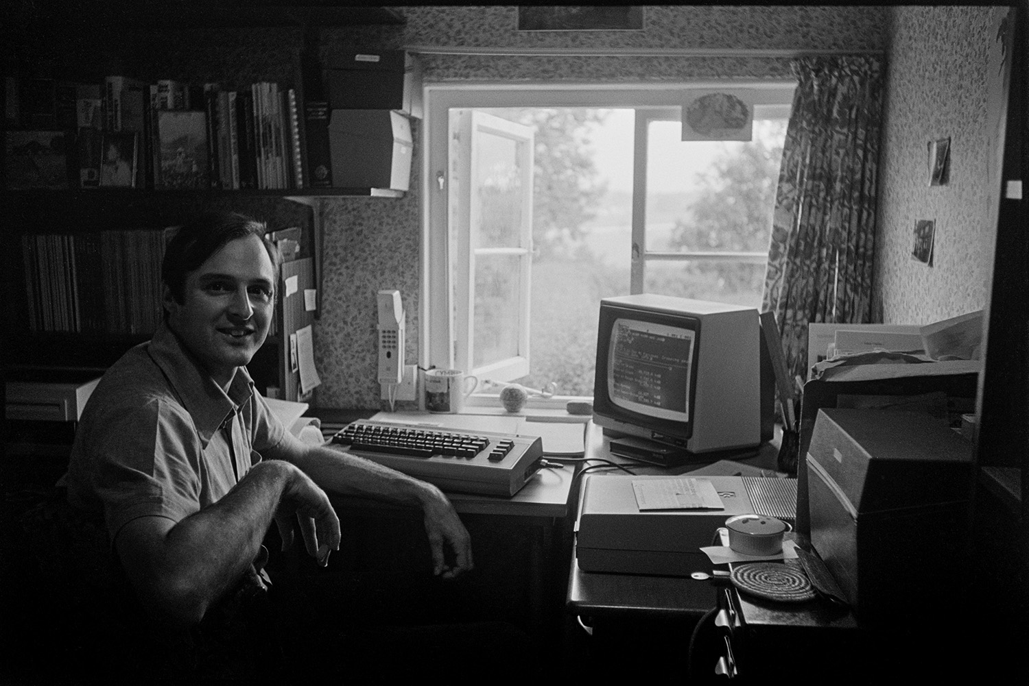 Man seated at desk with word processor. 
[A man sat at a desk with a computer in a house in Exbourne. Shelves with folders are next to an open window and a telephone is fixed to the wall. The walls are covered with patterned wallpaper.]