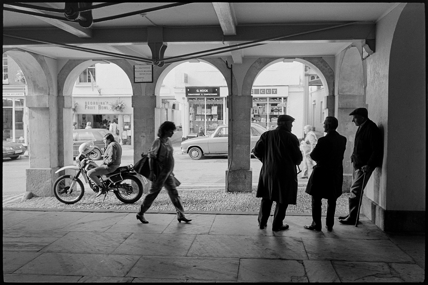 Men chatting under Town Hall arches, motorbike rider parked. 
[A woman walking past three men talking under the arches of the Town Hall in Torrington. A motorcyclist is also sat on his parked motorbike on the cobbles under the arches.]