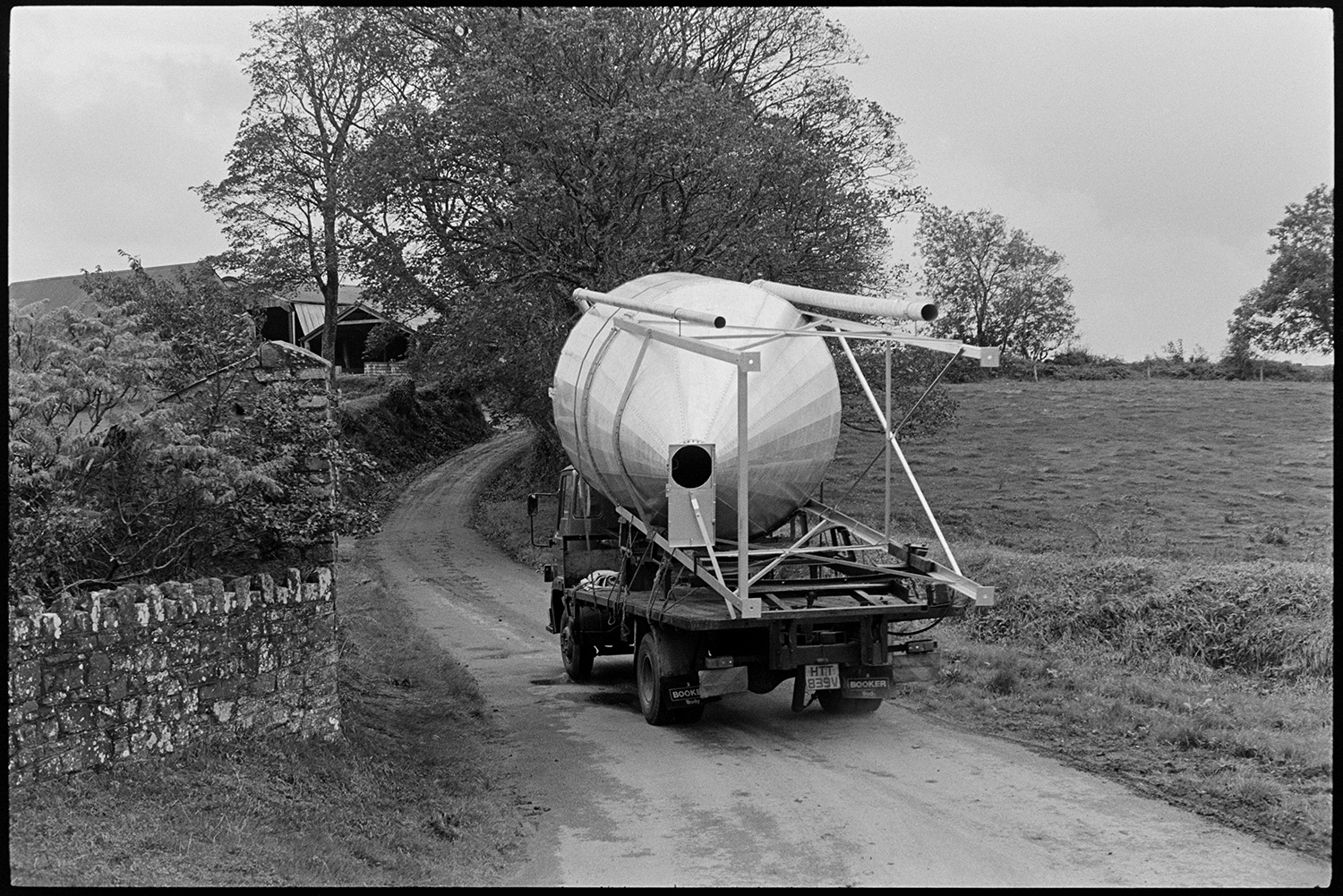 Lorry carrying large silo. 
[A lorry carrying a large silo up a lane to a farm at Harepath, Beaford. Farm buildings can be seen behind trees in the background.]