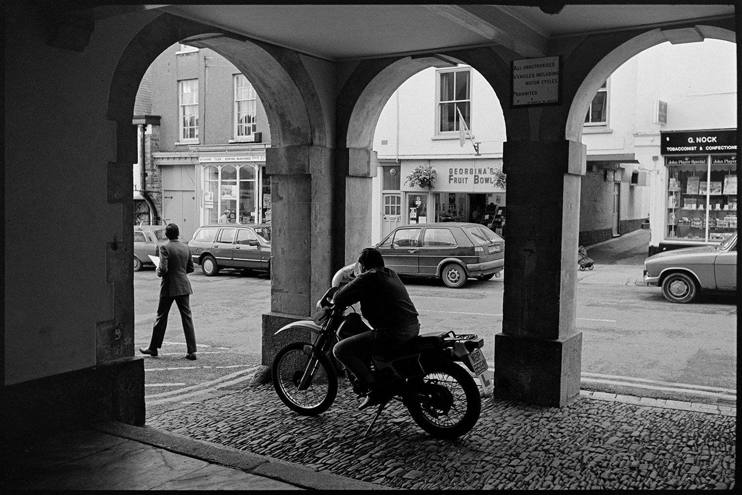 Men chatting under Town Hall arches, motorbike rider parked. 
[A motorcyclist sat on his parked motorbike on the cobbles under the arches of the Town Hall in Torrington. Parked cars and shop fronts can be seen on the opposite side of the street, including 'Georgina's Fruit Bowl' and 'G Nock Tobacconist and Confectioner'.]