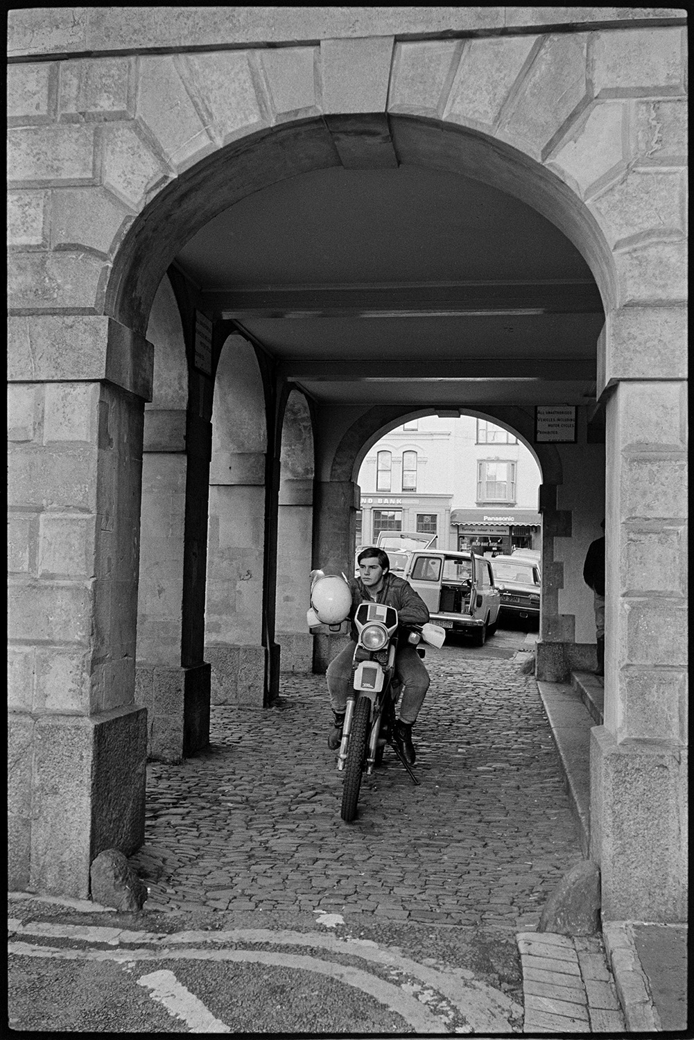 Men chatting under Town Hall arches, motorbike rider parked. 
[A motorcyclist sat on his parked motorbike on the cobbles under the arches of the Town Hall in Torrington. A parked van is visible in the background with shop fronts beyond.]