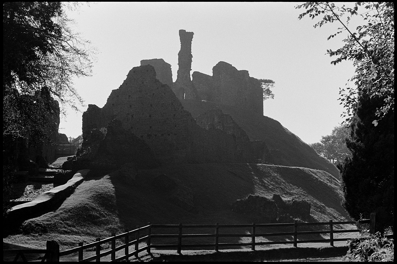 Ruined castle on mound. 
[The ruins of Okehampton Castle silhouetted against the sun. A modern wooden fence can be seen around the ruins.]