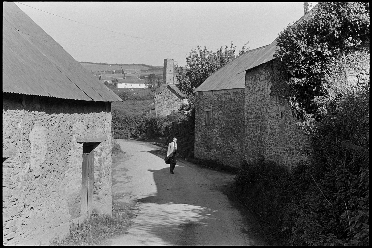 Doctor on his round in village street and visiting farm. 
[Doctor Richard Westacott walking along a road past stone and cob barns. A church tower can be seen in the background by other houses.]
