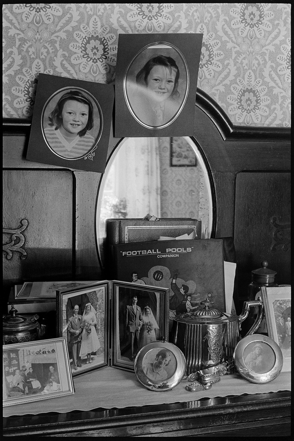 Side table with photographs and mirror. 
[A wooden side table in a house at Mariansleigh. Family photographs, a teapot and football pools folder are on display. A mirror is also set into the sideboard.]
