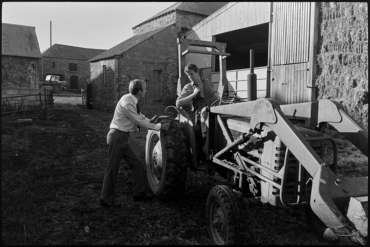 Doctor talking to friends on their farm. 
[Doctor Richard Westcott talking to Christopher Whinney who is sat on a tractor in the farmyard at Holdridge, North Molton. Barns, farm buildings and stacks of hay bales can be seen in the background.]