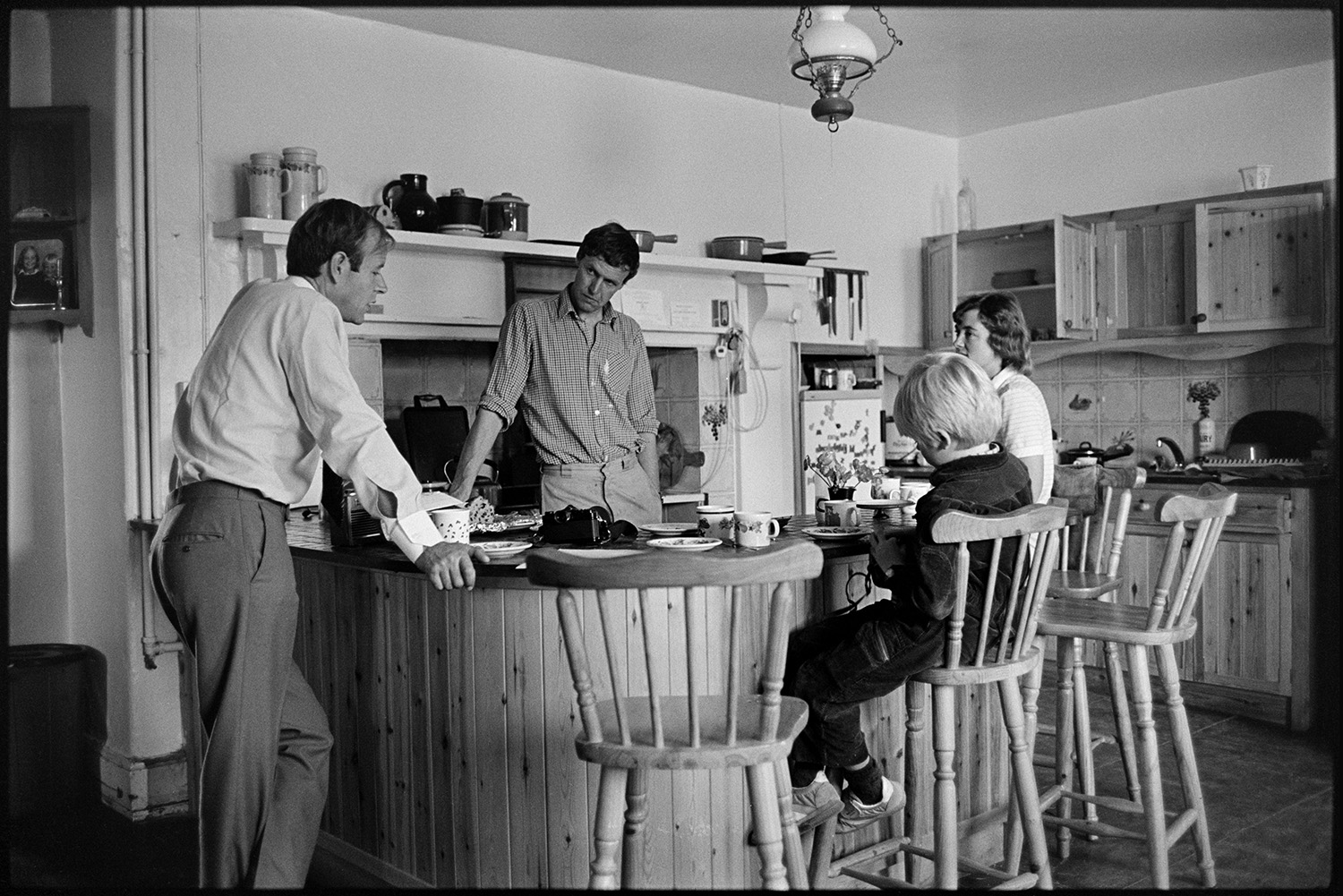 Doctor talking to friends on their farm. 
[Doctor Richard Westcott talking to Christopher Whinney and Anna Whinny in their kitchen at Holdridge, North Molton. Their child is sat at a breakfast bar in the kitchen. Various jars and saucepans are displayed above an Aga in the background.]