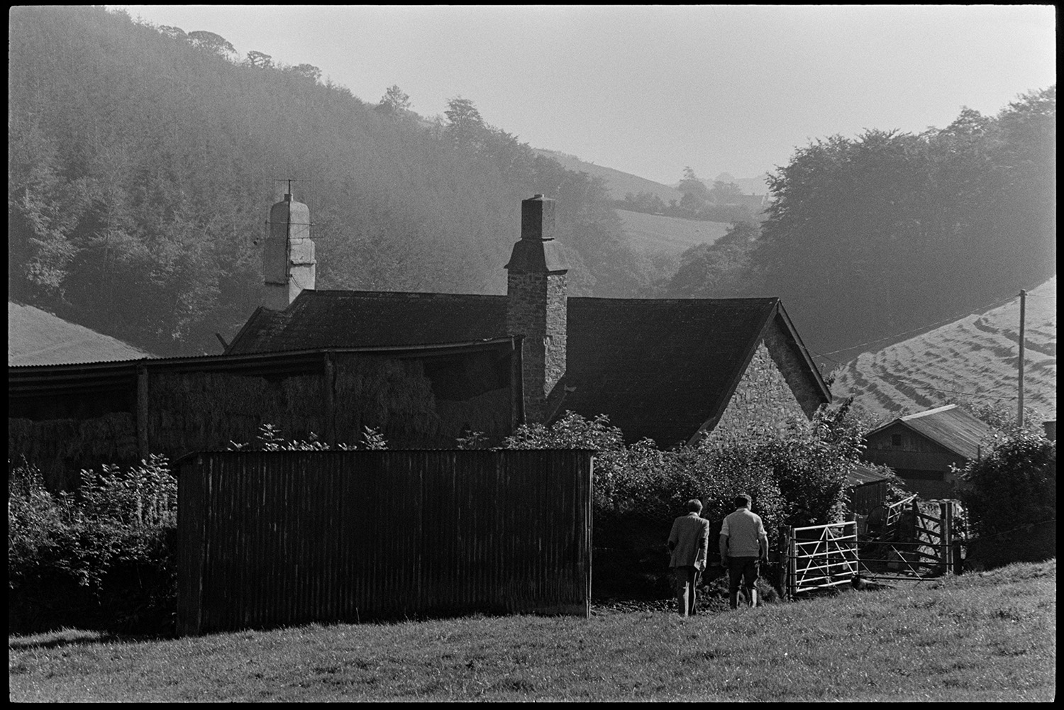 Doctor on his round visiting farm on moor. 
[Doctor Richard Westcott visiting a patient on a farm on Exmoor. He is walking with another man through a field towards the farmhouse. A barn with hay bales is also visible.]