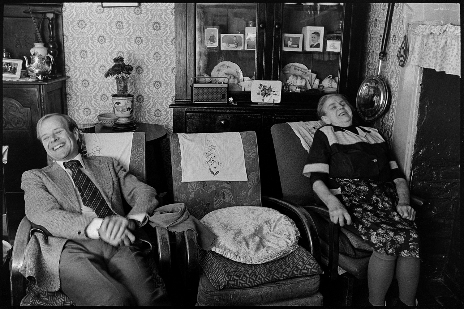 Doctor visiting woman patient, chatting in front of fireplace. Easy chairs and dresser, bellows. 
[Doctor Richard Westcott visiting a woman in her home at Mariansleigh. They are sat on chair in her living room and laughing. Photographs and china are displayed in a dresser behind them. A radio is also on the dresser. A fireplace and bed warmer hung on the wall are also visible. The wall is covered with patterned wallpaper.]