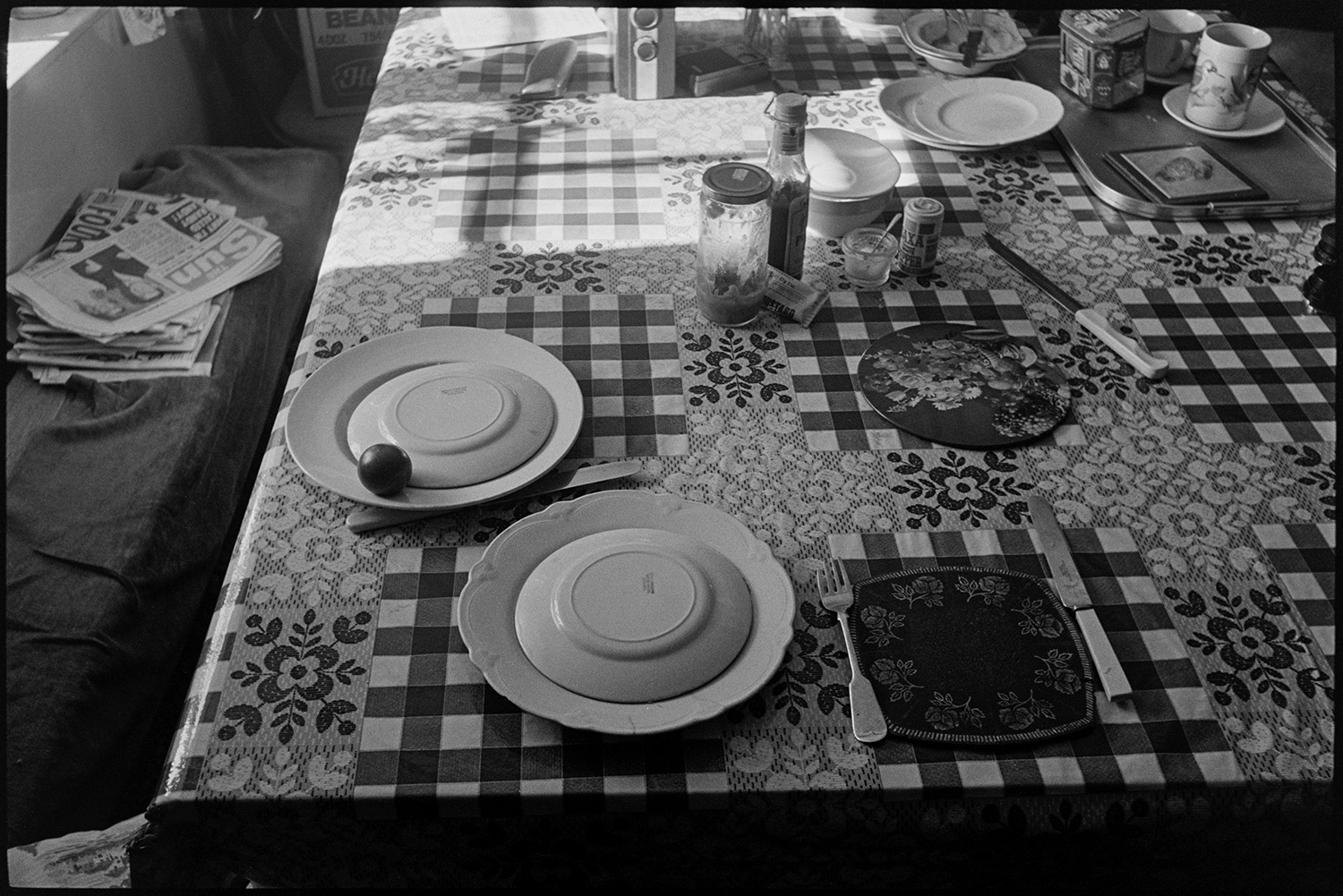 Cottage interior, sunlit table, tablecloth. 
[A sunlit kitchen table in a cottage at Mariansleigh. Plates, cups and jars are visible on the table. Two plates of food are covered. The table is covered with a tablecloth with a floral and lace design, and a small pile of newspapers is on the settle next to the table.]