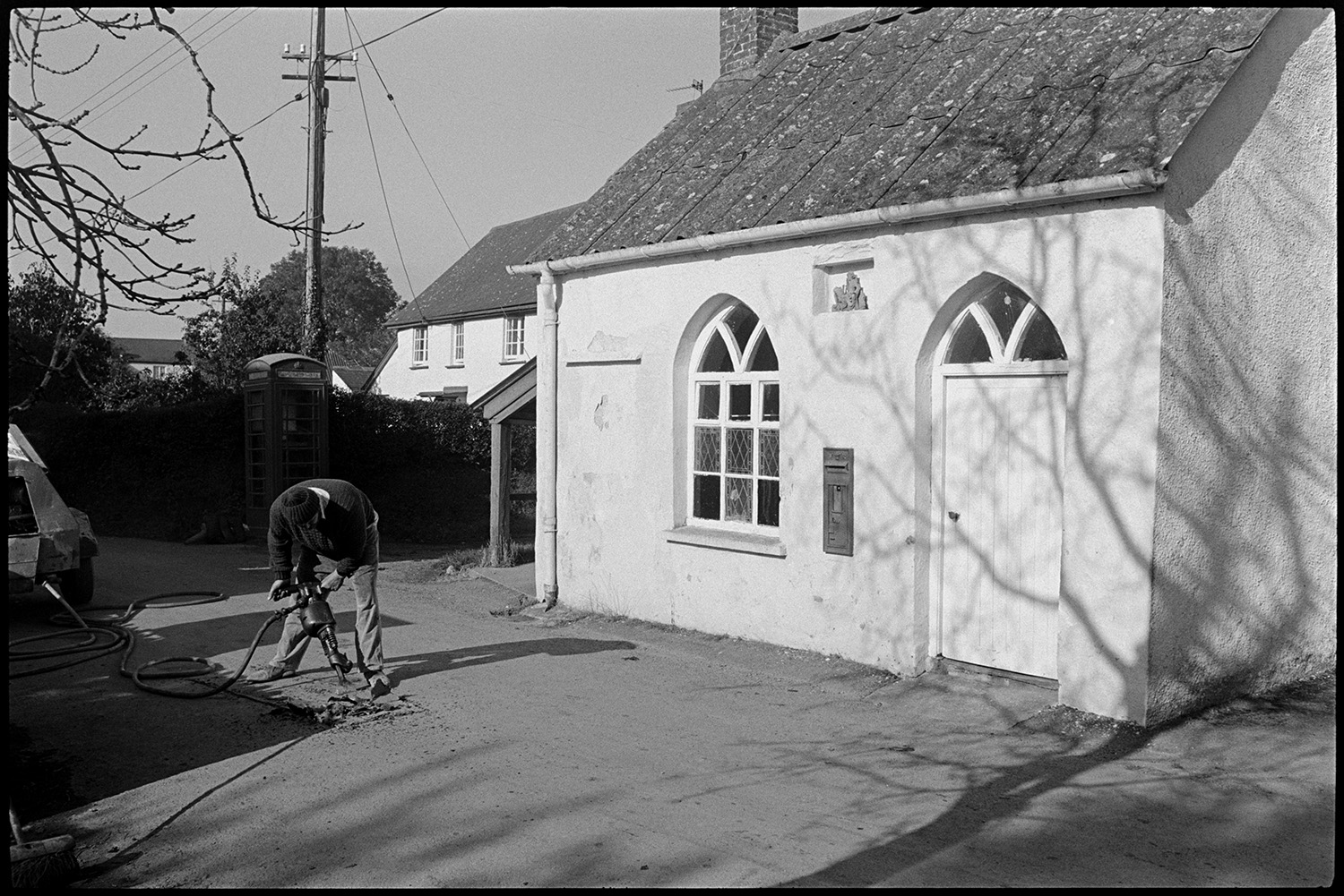 Man drilling hole in road in front of Chapel. 
[A man using a pneumatic drill to dig a hole in the road in front of a chapel and telephone box at Mariansleigh. A post box is set into the wall of the chapel.]
