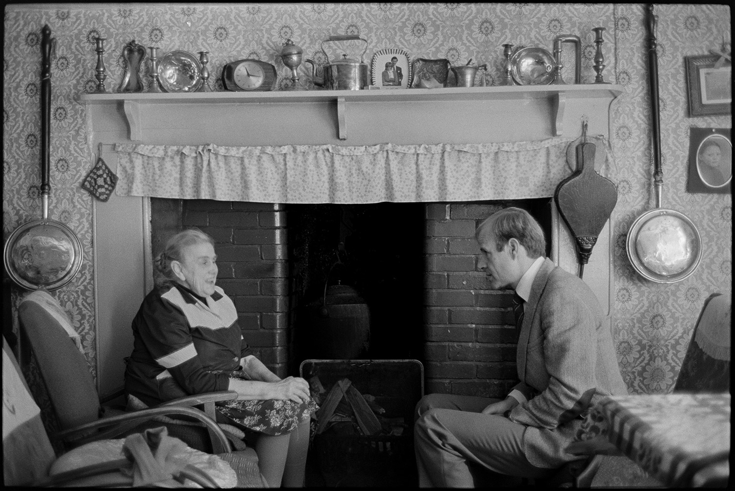 Doctor visiting woman patient, chatting in front of fireplace. Easy chairs and dresser, bellows. 
[Doctor Richard Westcott visiting a patient in her home at Mariansleigh. They are sat on chairs in the living room in front of a fireplace. A display of ornaments, candlesticks and a kettle is on the mantelpiece and a pair of bellows are hung by the fire. The wall is covered with patterned wallpaper and two bed warmers are hung either side of the fireplace.]