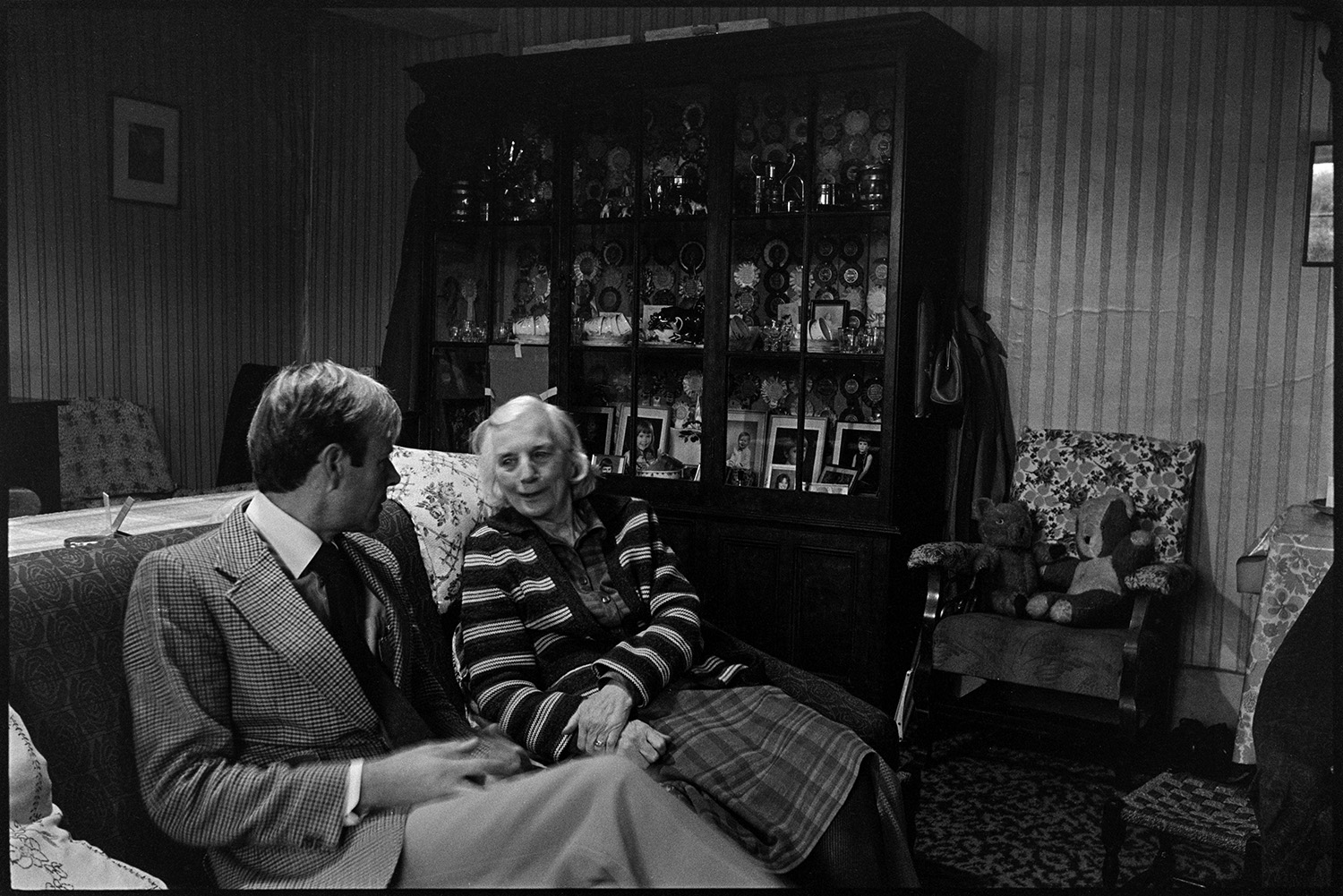 Doctor on rounds talking to woman patient in her sitting room. Fireplace, dresser. 
[Doctor Richard Westcott talking to Mrs Balment sat on a sofa in her living room in her house at Stoodleigh, Brayford. A display cabinet with photographs and china can be seen in the background.]