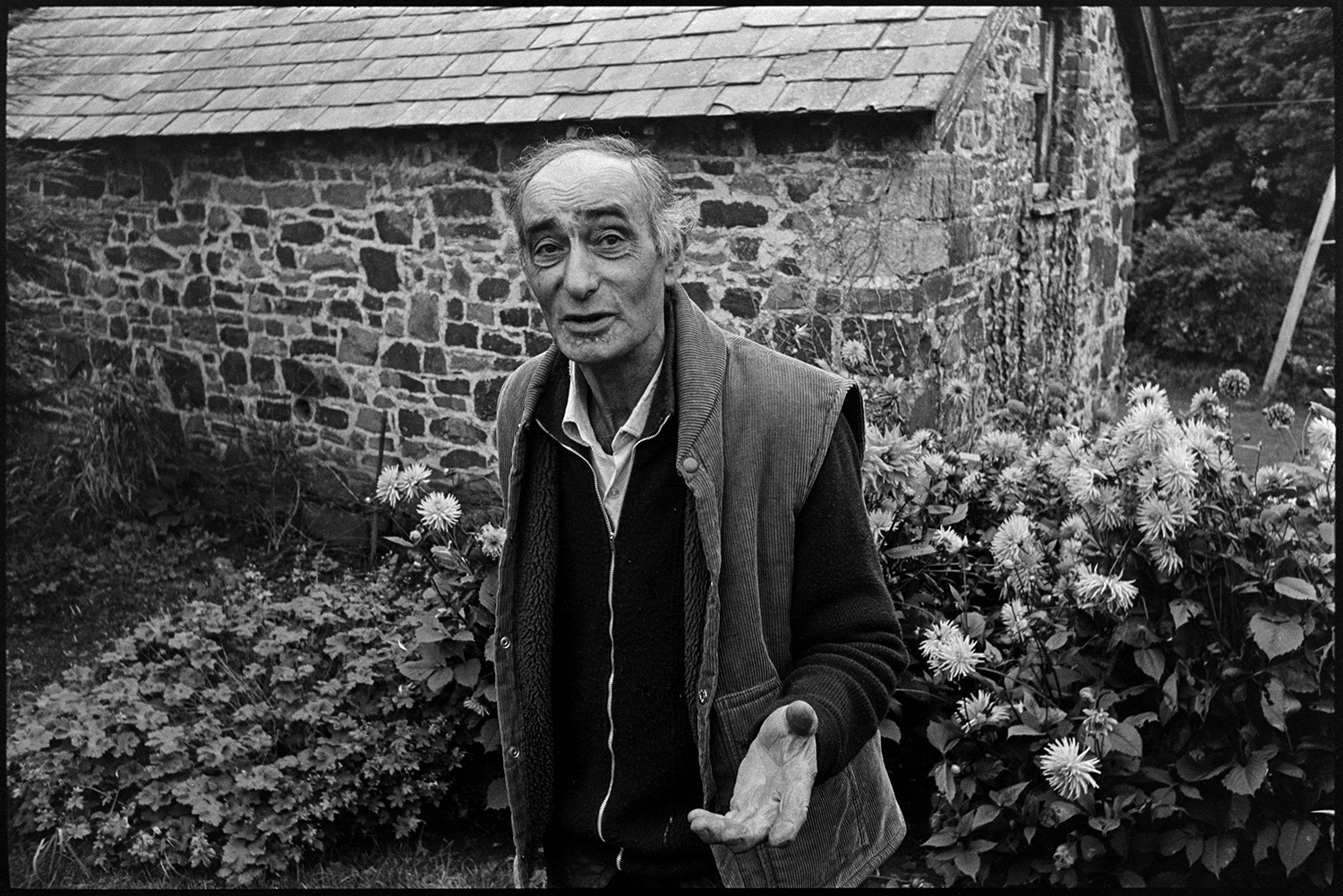 Doctor on rounds talking to man patient outside his house looking at his flowers. 
[Mr Bolzone talking to Doctor Richard Westcott (not pictured in the image) by flowers and a stone and slate building in his garden.]
