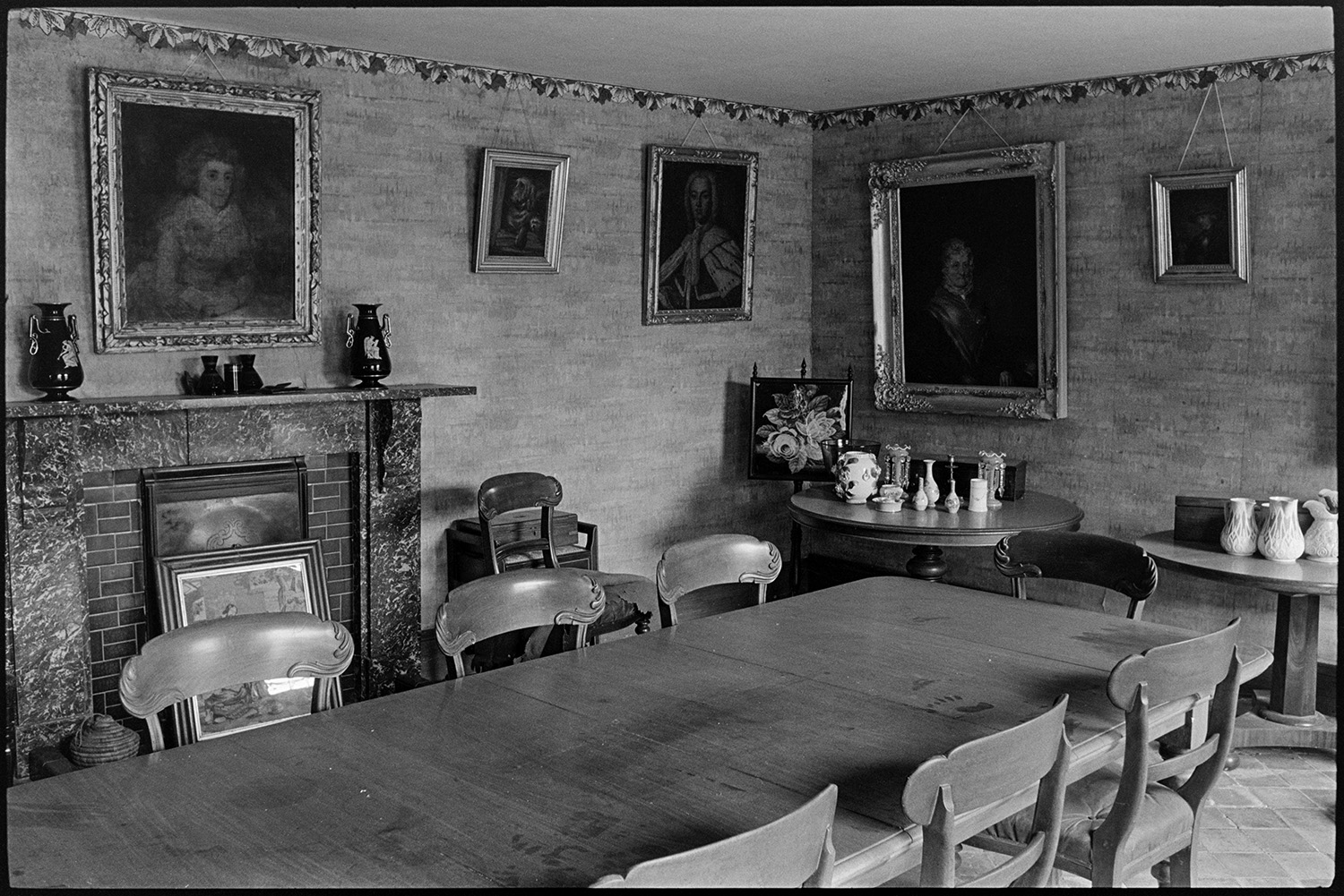 Dining room of large farmhouse. Furniture, table, chairs, china, pictures. 
[The dining room in the farmhouse at Narracott Farm, South Molton. A large table with chairs, a fireplace, side table with jugs and pictures hung on the wall are visible.]