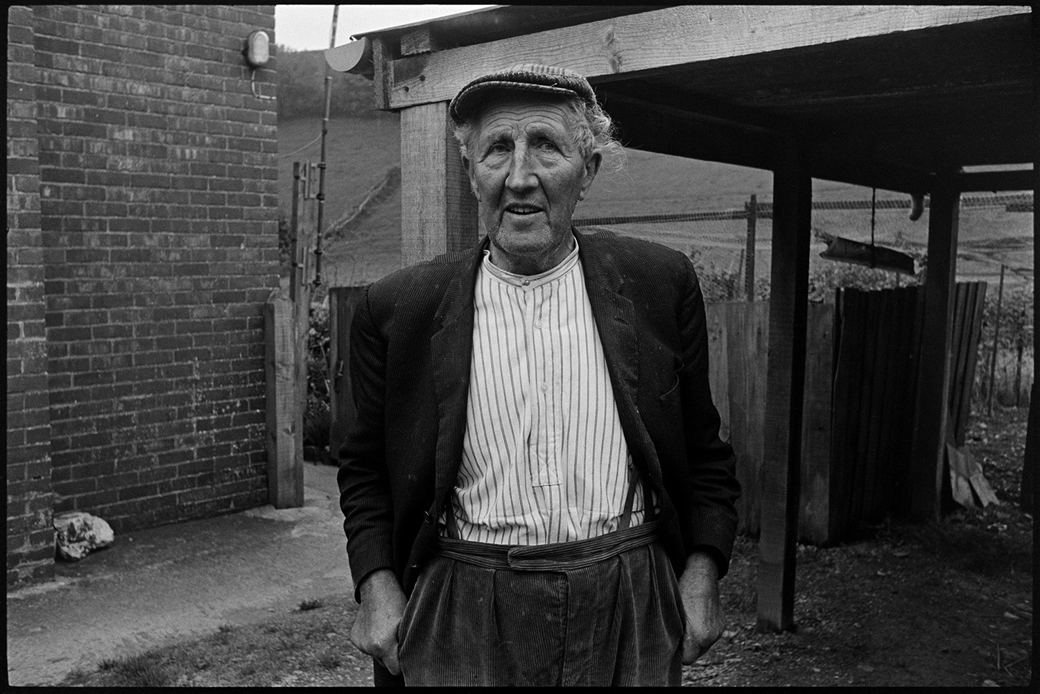 Doctor on rounds talking to man patient outside his house looking at his vegetable patch. 
[Edward Cotsford stood outside his house talking to Doctor Richard Westcott (not pictured in the image). He is stood by a wooden open barn and his vegetable garden is partially visible in the background.]