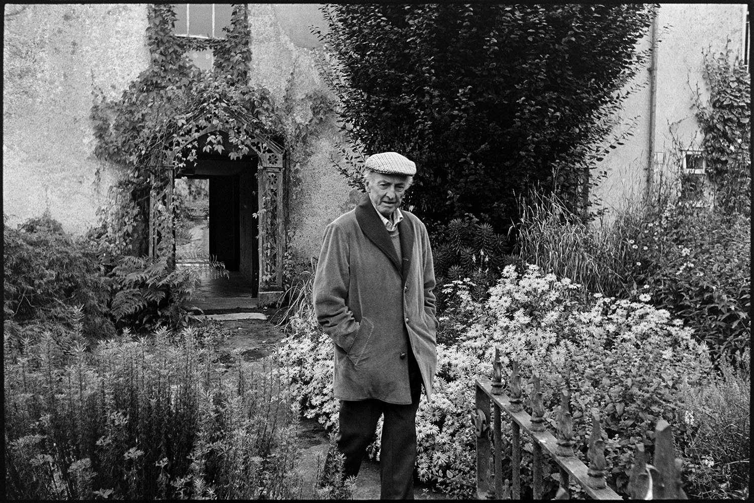 Doctor on rounds talking to man patient in kitchen with panelling  and table drawers. 
[John Huxtable walking through a garden with flowers at Narracott, South Molton. A plant is growing around the carved entrance to the building in the background.]