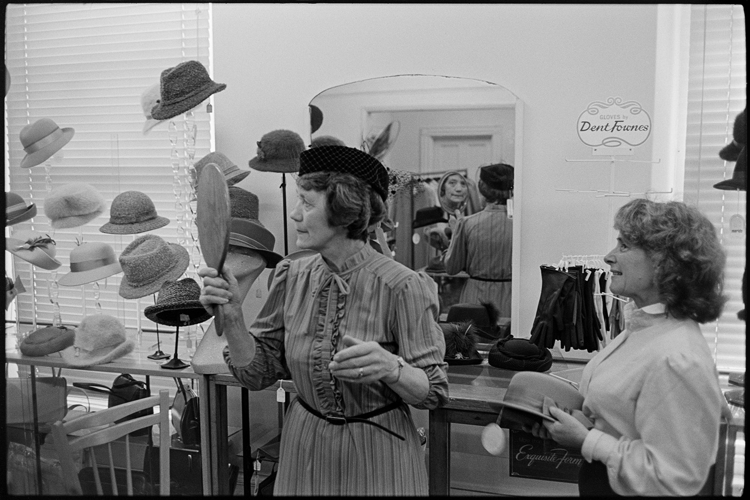 Shop assistant helping woman choose a hat in hat department of clothes shop. Hat display. 
[A shop assistant in Trapnells clothes shop in Bideford High Street, helping a woman choose a hat. The customer is holding a mirror and looking at the hat she is trying on. A hat display can be seen in the background.]