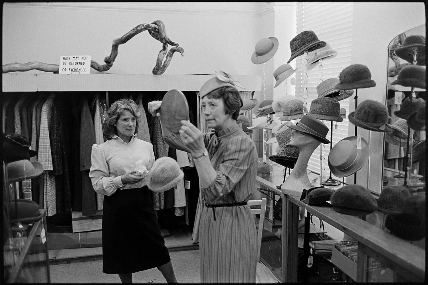 Shop assistant helping woman choose a hat in hat department of clothes shop. Hat display. 
[A shop assistant in Trapnells clothes shop, in Bideford High Street, helping a woman choose a hat. The customer is holding a mirror and looking at the hat she is trying on. A hat display and clothes rail can be seen in the background.]