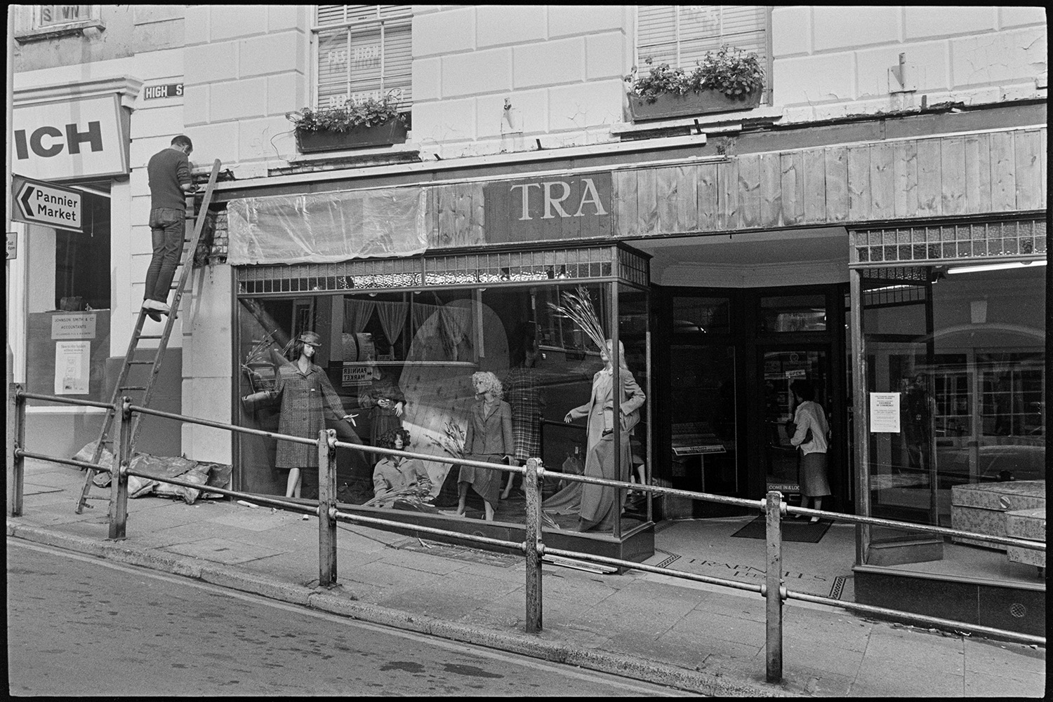 The front of Trapnells clothes shop being renovated shortly before the shop closed, in Bideford High Street. A man is up a ladder working on the shop sign. Mannequins modelling clothes can be seen in the shop front window.