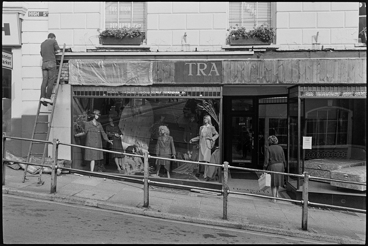 Front of clothes shop being repaired shortly before it closed. 
[The front of Trapnells clothes shop being renovated shortly before the shop closed, in Bideford High Street. A man is up a ladder working on the shop sign. Mannequins modelling clothes can be seen in the shop front window and a woman is walking into the shop.]