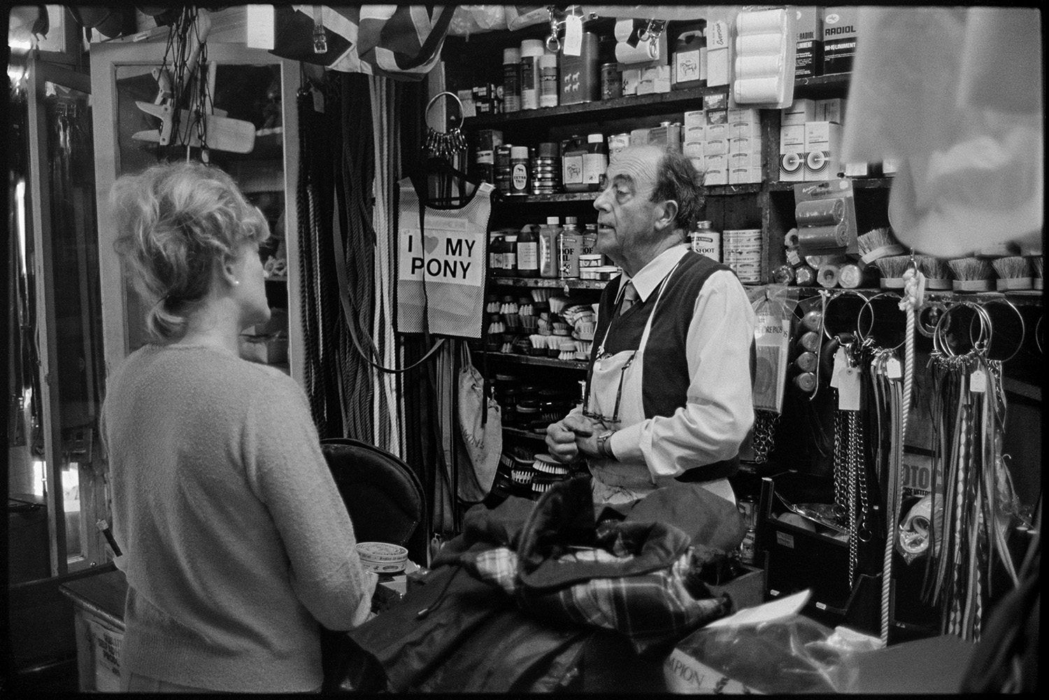 Saddlers shop, proprietor and customer. <br />
[Derek Johns serving a woman in his saddlers shop in Buttgarden Street, Bideford. Various good are on display behind the counter, including brushes, bottles, rollers and chains.]