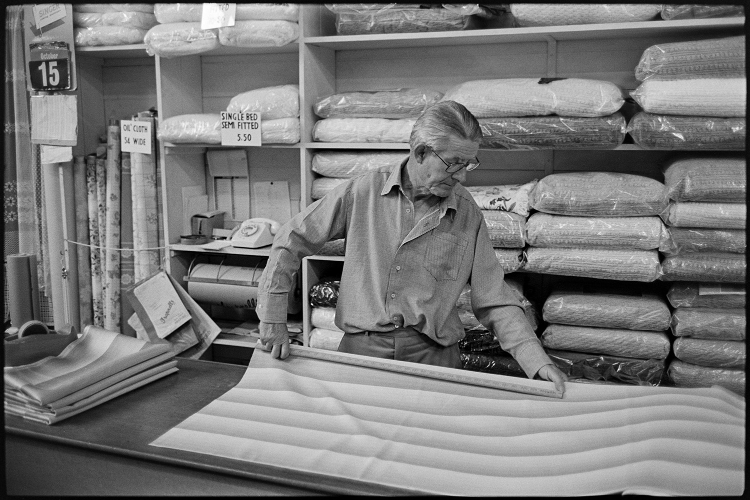Proprietor of clothes shop, measuring cloth and talking to customer. 
[Mr Trapnell measuring material in his clothes shop, Trapnells, in Bideford High Street. Rolls of material and bedding can be seen on the shelves in the background.]